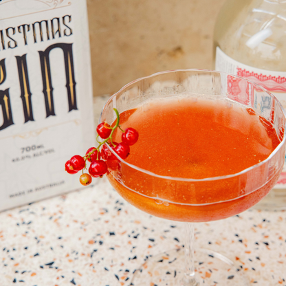 Cocktail with red berries in a glass next to a bottle of Christmas Gin on a speckled surface.