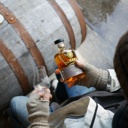 A person wearing knit gloves holds a bottle of McHenry Lightly Peated Whisky and a clear glass next to a large wooden barrel.