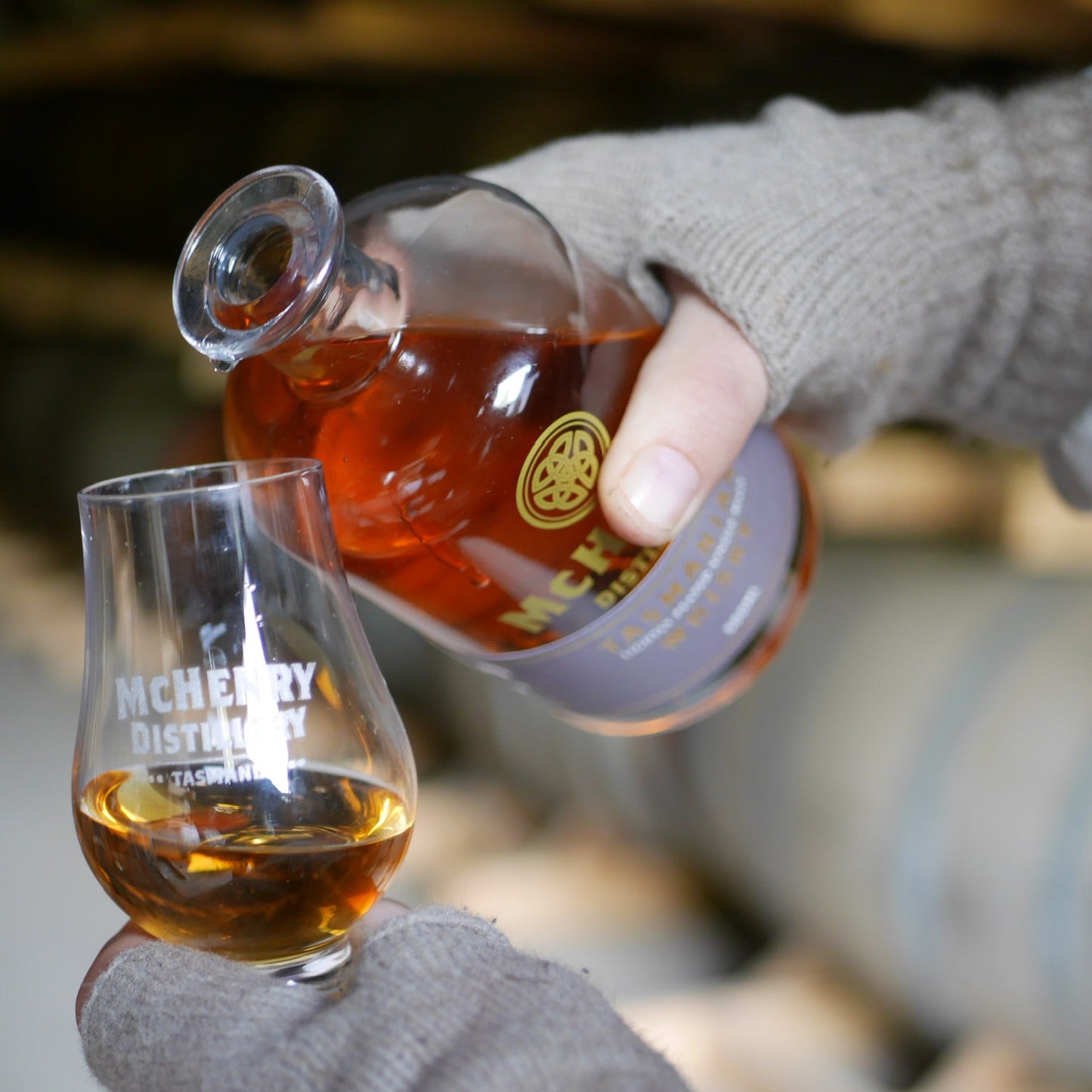 A person wearing gray knit gloves pours whisky  a clear glass snifter cup with white text, in a blurred indoor setting.