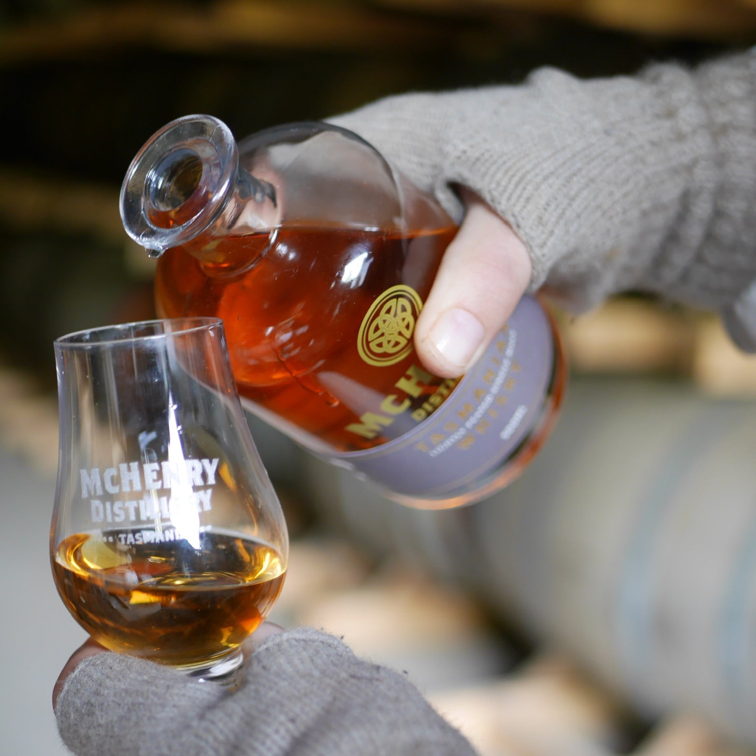 A person wearing gray knit gloves pours whisky  a clear glass snifter cup with white text, in a blurred indoor setting.