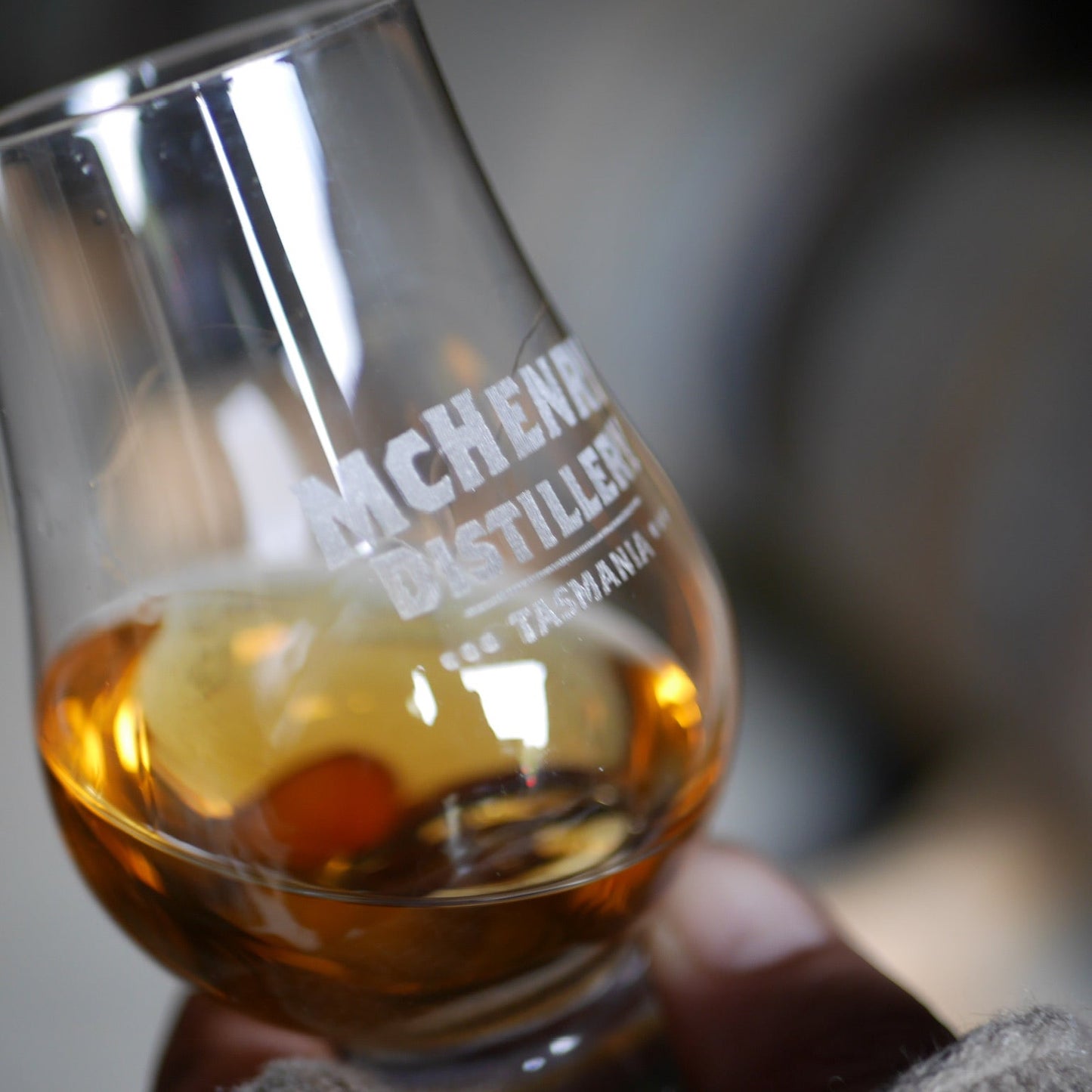 A close-up of a hand holding a clear glass with whisky, featuring etched text that reads 'MCHENRY DISTILLERY TASMANIA'