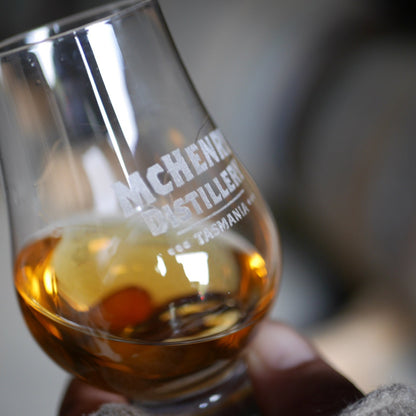 A close-up of a hand holding a clear glass with whisky, featuring etched text that reads 'MCHENRY DISTILLERY TASMANIA'