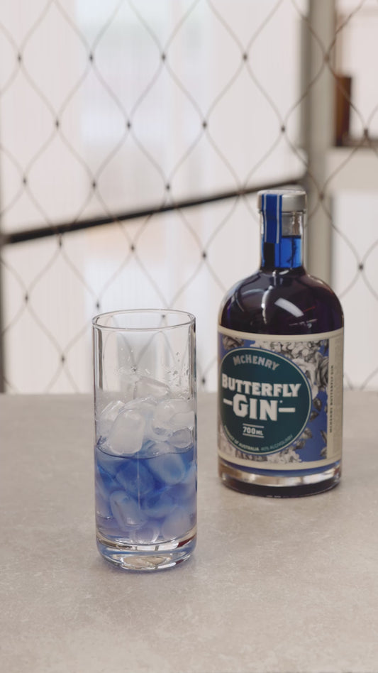 Pouring tonic water into a glass of Butterfly Pea Flower Gin over ice, causing a vibrant colour change reaction, with gin bottle on white background.